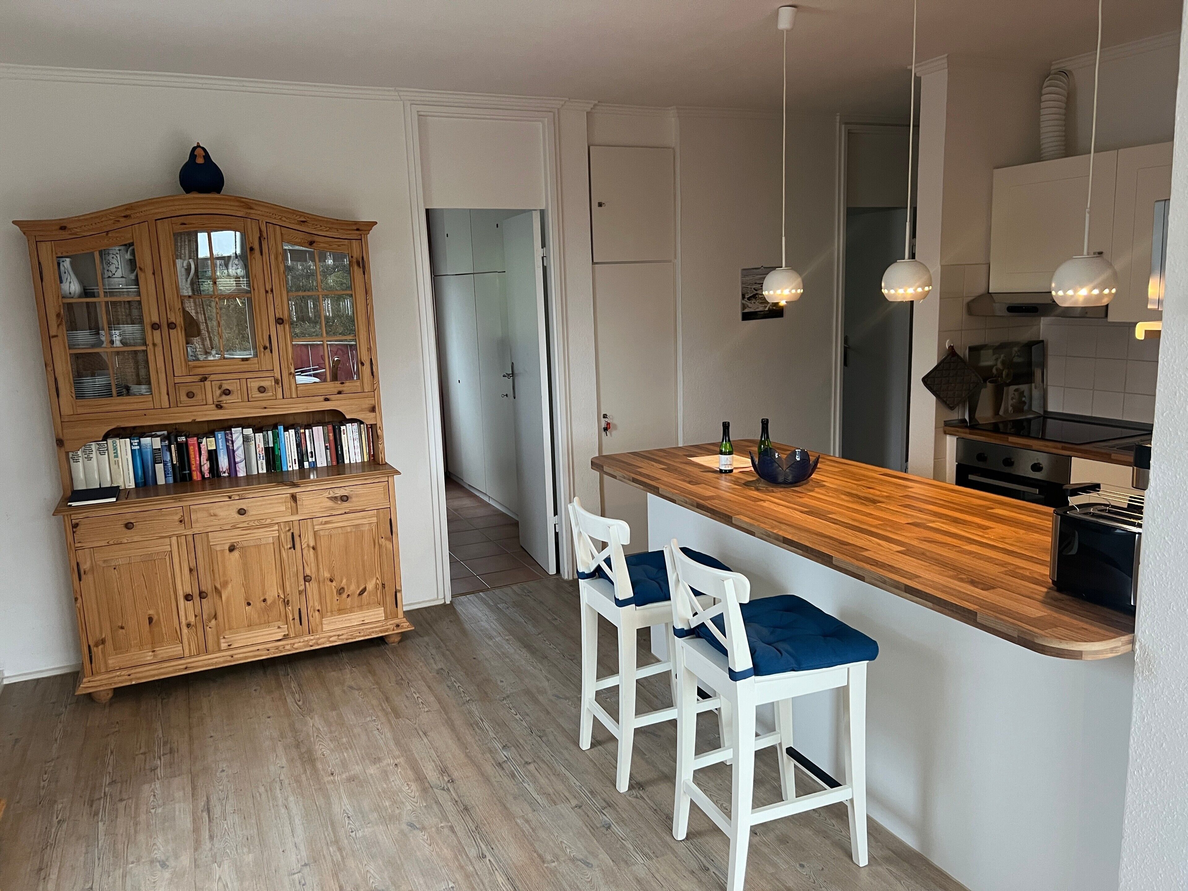 Une cuisine avec des meubles de rangement en bois, deux tabourets de bar, un plan de travail en bois et des luminaires suspendus.