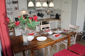 A dining area with a wooden table set for four, white chairs, a vase with red flowers, and a kitchen in the background.