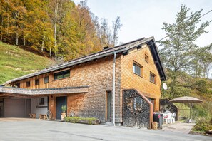 A modern wooden house with a garage, outdoor seating, and a stone wall. - Holiday Home 'Am Berg' with Mountain View, Terrace, Sauna and Wi-Fi (Hirschau)
