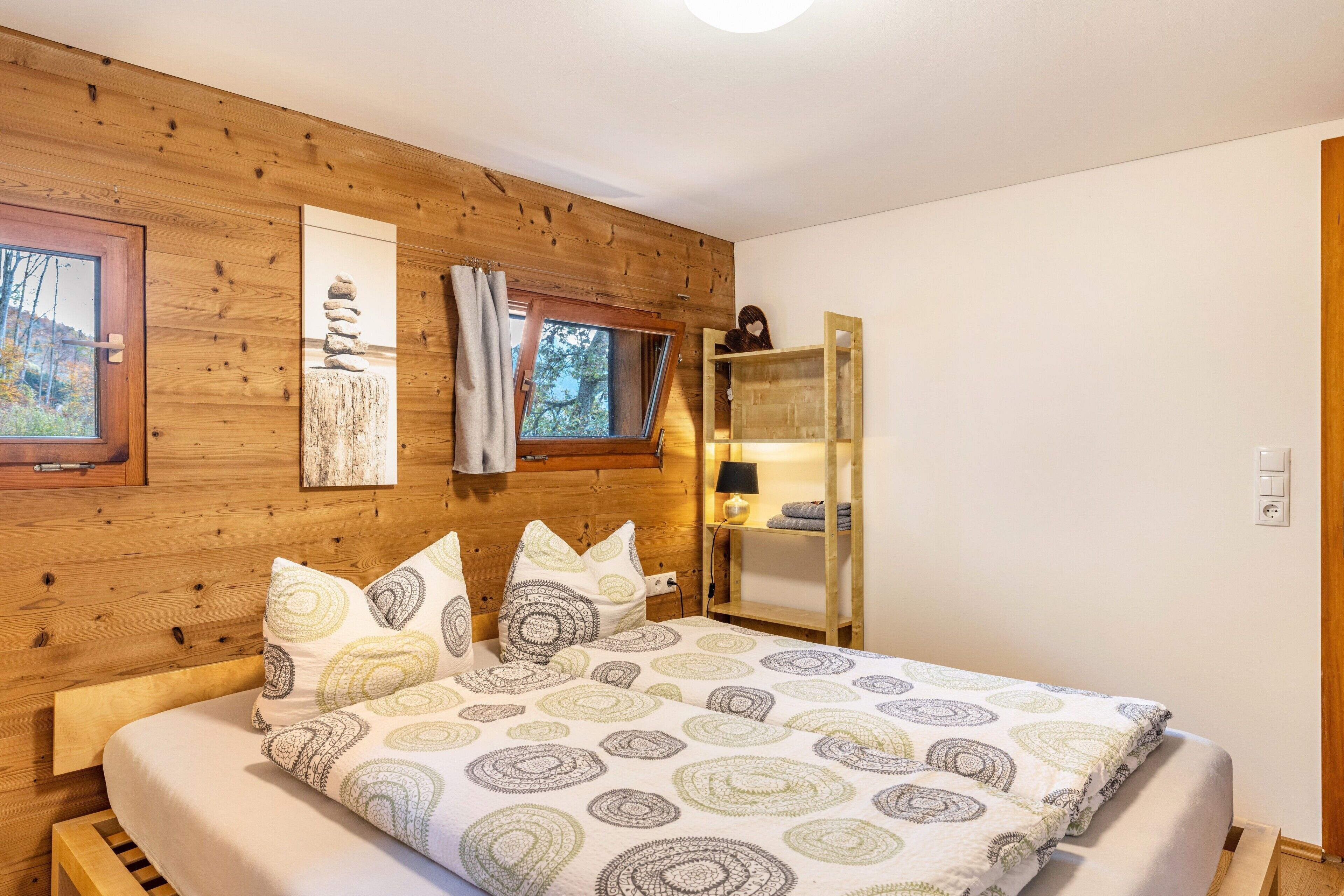 A wooden-paneled bedroom with a bed, a window, a shelf with items, and a wall-mounted lamp.