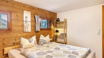 A wooden-paneled bedroom with a bed, a window, a shelf with items, and a wall-mounted lamp.