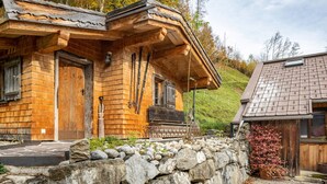 A wooden cabin with a stone wall in front, a wooden door, and a window with a wooden shutter.