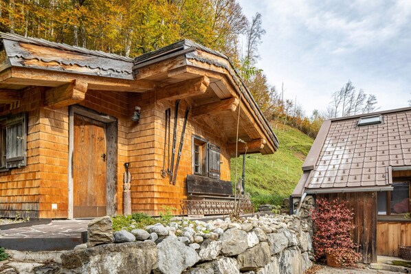 A wooden cabin with a stone wall in front, a wooden door, and a window with a wooden shutter. - Holiday Home 'Am Berg' with Mountain View, Terrace, Sauna and Wi-Fi (Hirschau)