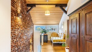 A narrow hallway with a brick wall, wooden ceiling beams, a bed with yellow bedding, a potted plant, and a wooden wardrobe.