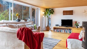 A modern living room with a sofa, a red chair, a TV, a bookshelf, and a large window with a view of trees.