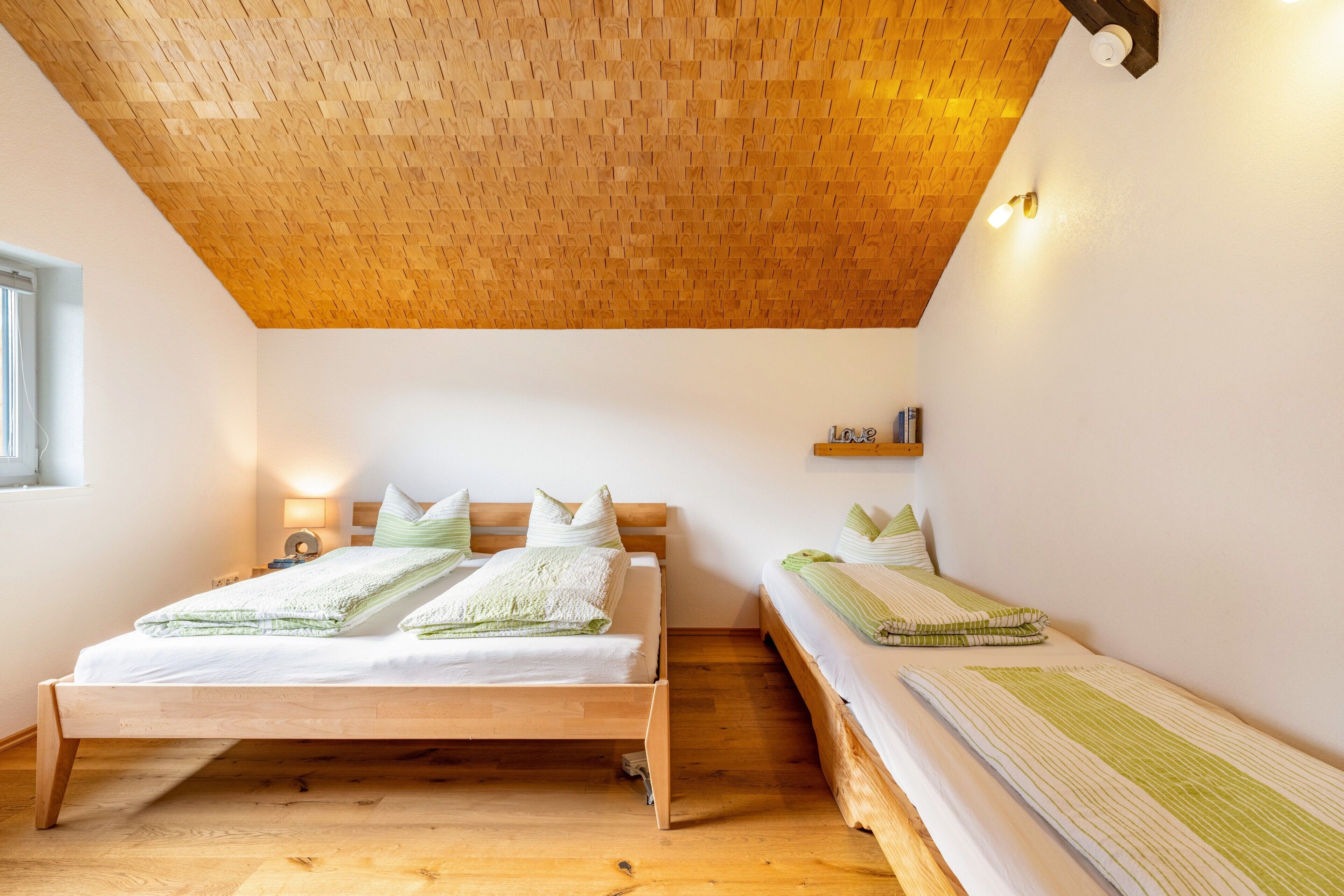 Une pièce avec un plafond en bois, un lit avec une literie verte, une table de chevet avec une lampe et une petite étagère remplie de livres.
