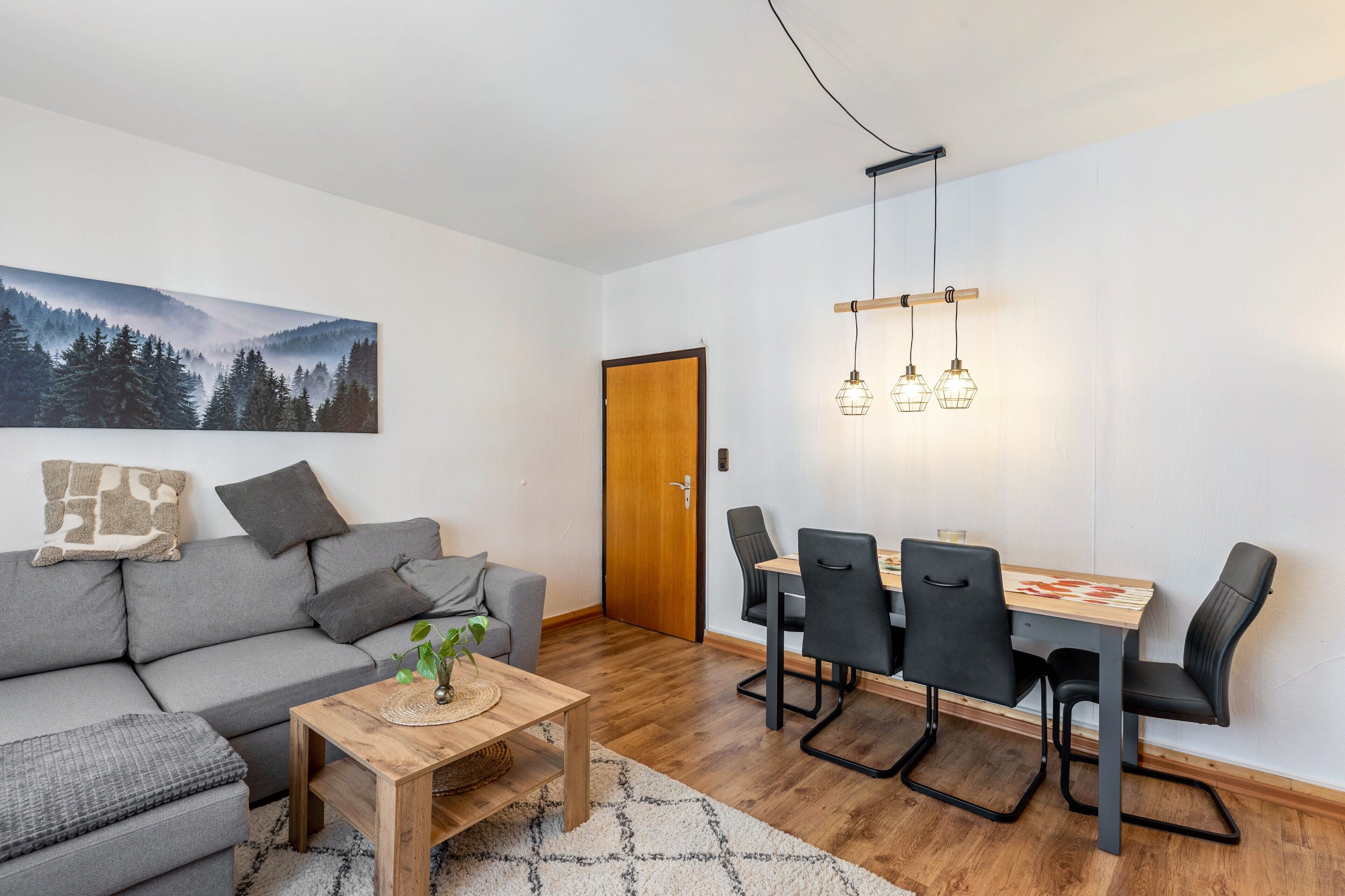 A modern living room with a gray sofa, a wooden coffee table, a dining table with four chairs, and a painting of a forest on the wall.