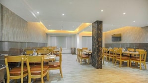 A spacious dining area with multiple wooden tables and chairs, a central pillar, and a television mounted on the wall. - Aangan Premium Shimla (Shimla)