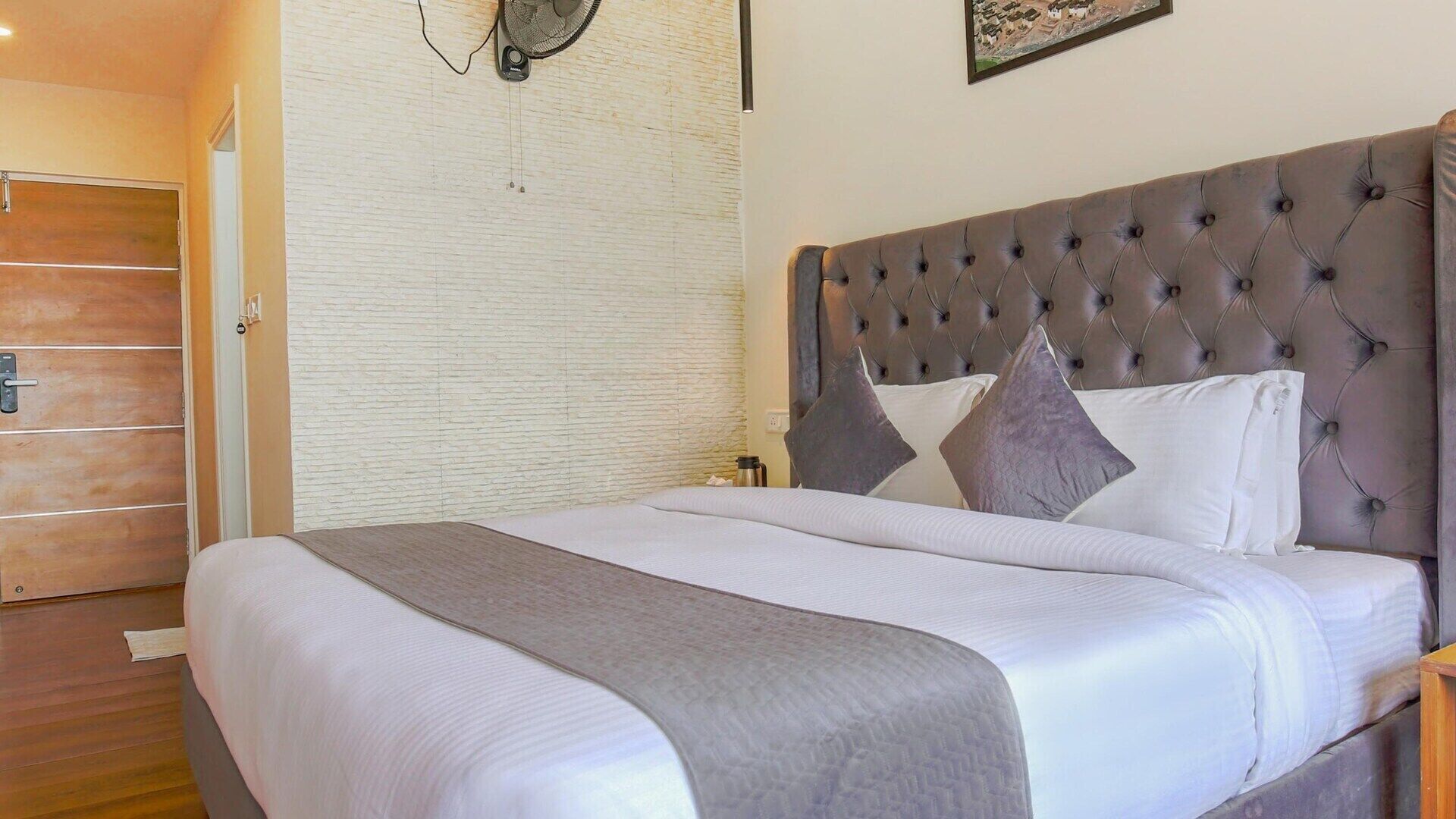 A neatly made bed with a tufted headboard, a bedside table, and a wall-mounted fan.