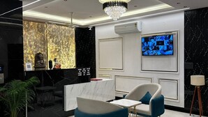 A modern reception area with a marble reception desk, a chandelier, a TV mounted on the wall, and a seating area with a blue sofa and a white armchair. - The Crown Prince (Greater Noida)