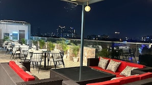 An outdoor seating area with wicker furniture and red cushions, overlooking a cityscape at night. - The Crown Prince (Greater Noida)
