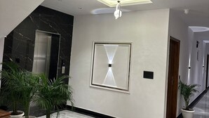 A modern hallway with marble flooring, potted plants, a central light fixture, and a ceiling with recessed lighting. - The Crown Prince (Greater Noida)
