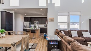 A modern kitchen with dark cabinets, a central island, and a dining area with a wooden table and chairs. - West Jordan Modern Home | Fenced Yard, New Build, Sleeps 7! (West Jordan)