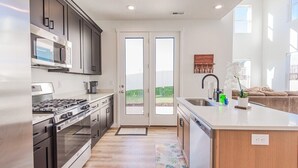 A modern kitchen with stainless steel appliances, a large island, and a view through glass doors to the outdoors. - West Jordan Modern Home | Fenced Yard, New Build, Sleeps 7! (West Jordan)