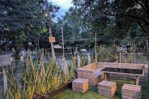 Un espace extérieur aménagé avec des bancs et des tables en bois, entouré de hautes herbes et d’arbres.
