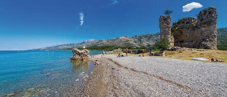 A pebbled beach with clear water, rocky ruins, and mountains in the background.