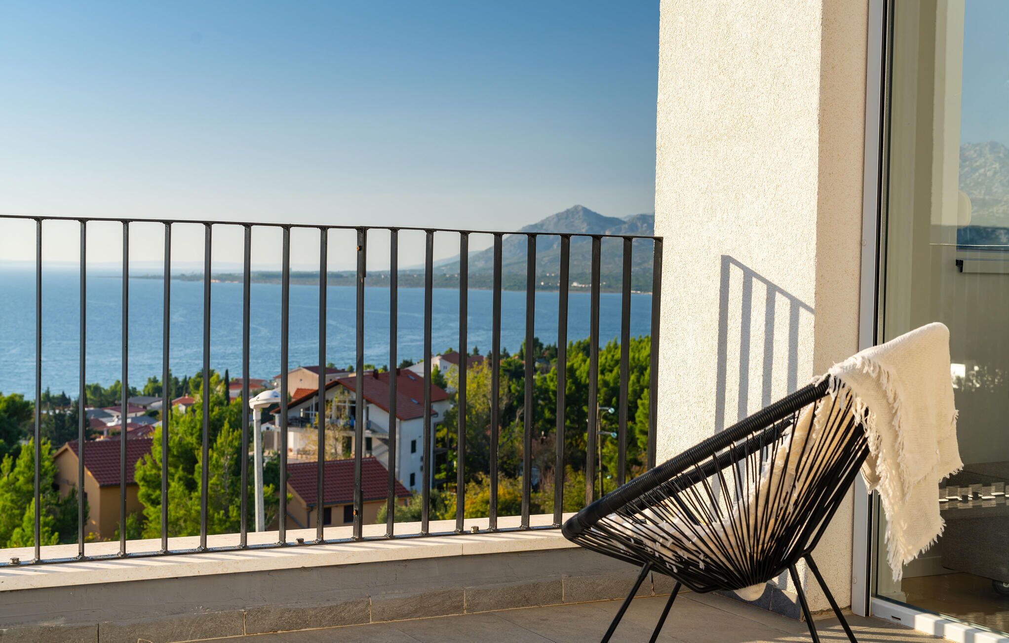 A balcony with a view of the sea and houses, a black chair, and a white towel.