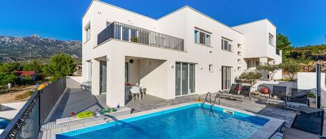 A modern two-story house with a swimming pool, outdoor seating, and a view of mountains.
