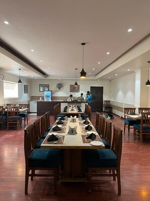 A dining area with a long table set for multiple guests, wooden chairs, and a counter with staff in the background. - Hotel Vasu International  (Phagwara)