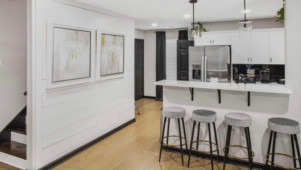 Een moderne keuken met witte kastjes, een centraal eiland met krukken, en twee abstracte schilderijen aan de muur.