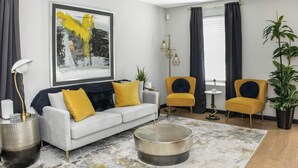 Een moderne woonkamer met een grijze bank, gele kussens, een ronde salontafel en twee gele fauteuils. Aan de muur hangt een groot abstract schilderij en staat een vloerlamp.