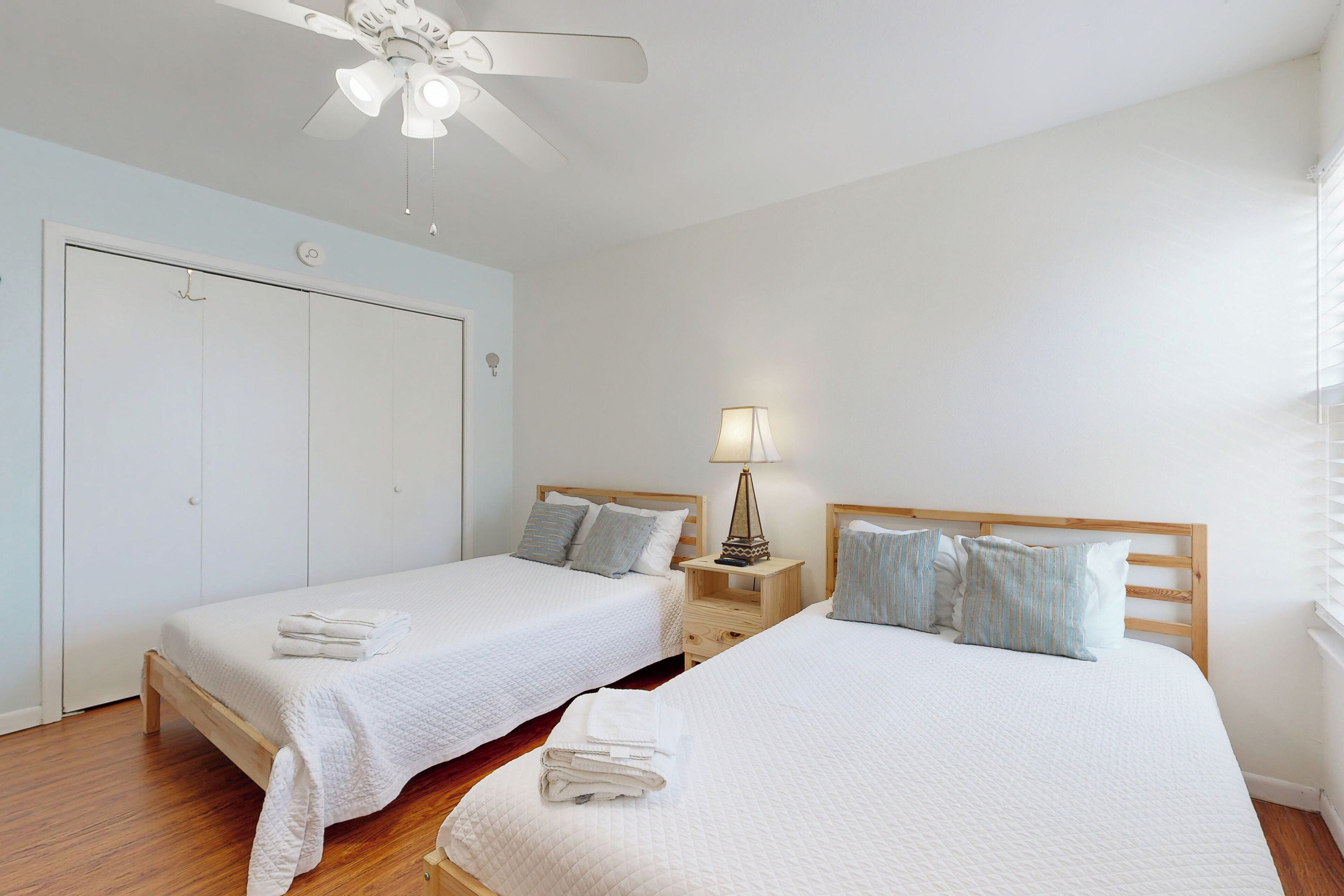 A hotel room with two single beds, a ceiling fan, a bedside table with a lamp, and a wardrobe.