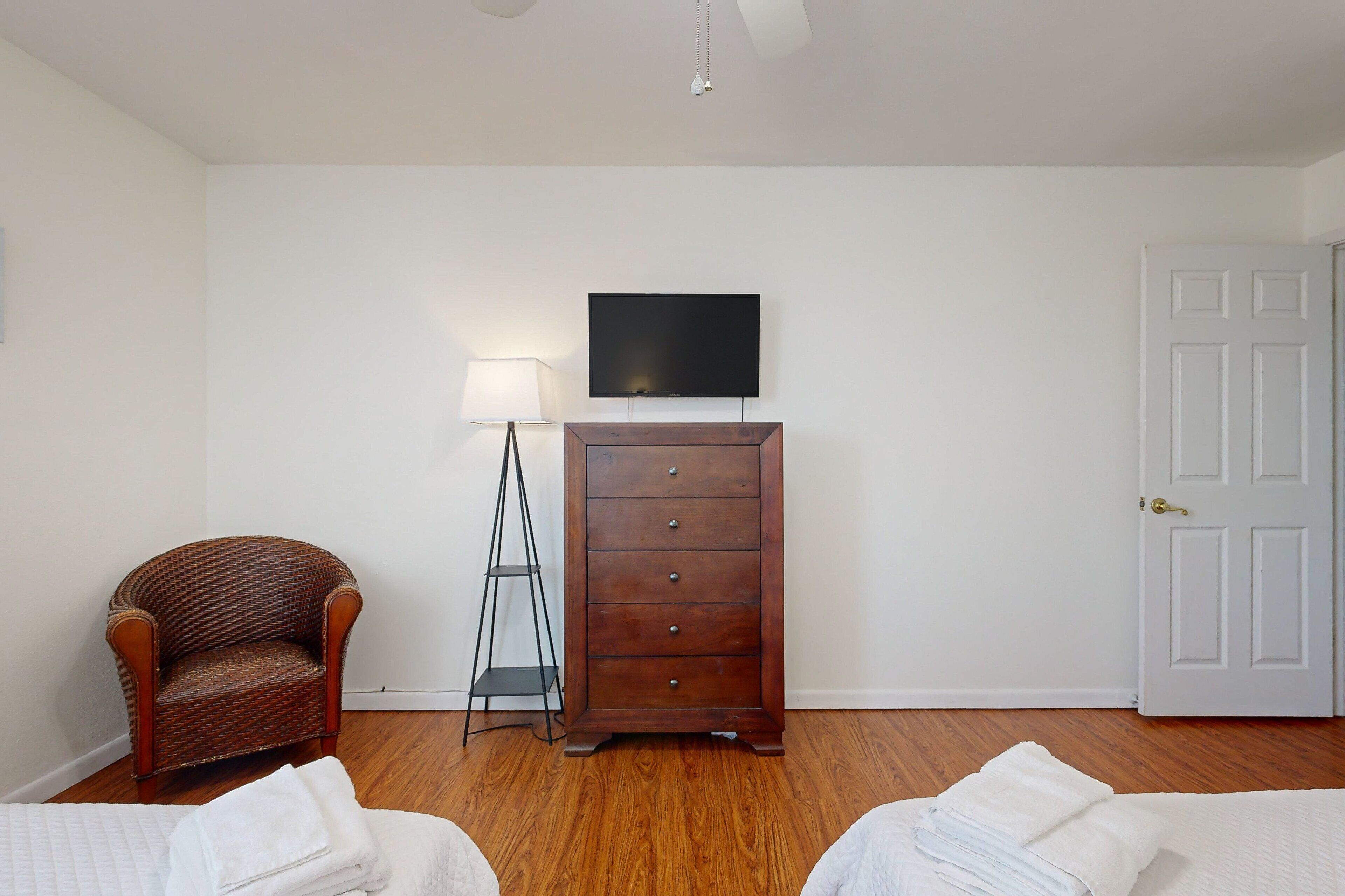 Une pièce comprenant une commode en bois, un téléviseur fixé au mur, une chaise et un lit avec une literie blanche.