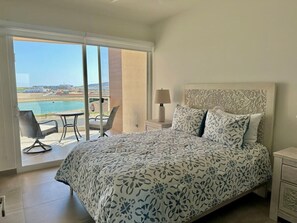 Relax in this serene room with a large bed, a window offering a view of a body of water and distant landscape, and a comfortable chair and table nearby.