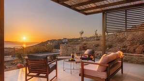 Um terraço com móveis de madeira, uma mesa com uma garrafa e vista para o pôr do sol sobre o mar.
