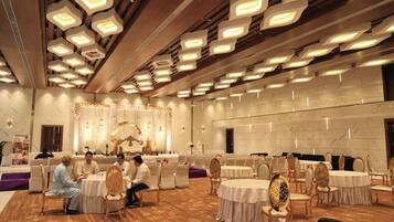 A banquet hall with round tables and chairs, a stage with a floral arrangement, and a ceiling with recessed lighting.