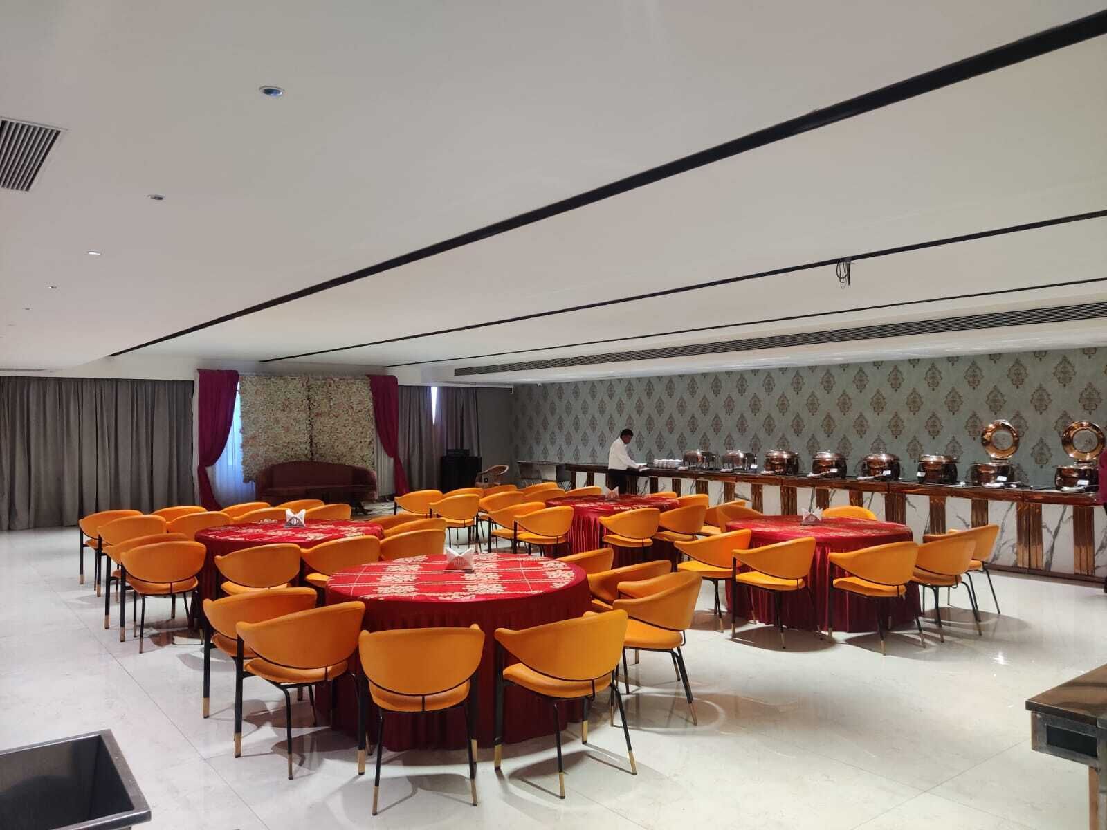 A conference room with round tables and orange chairs, a stage with a red curtain, and a buffet line with dishes.