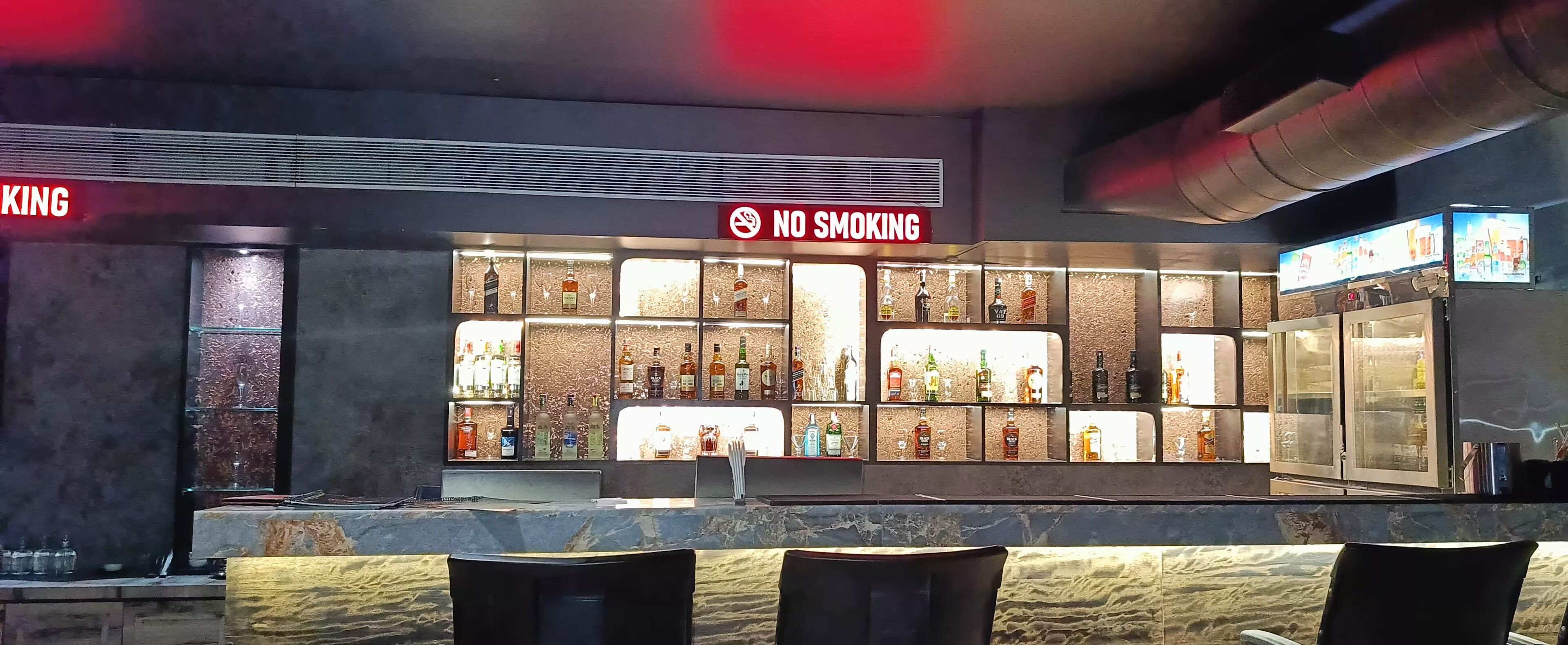 A bar area with a 'No Smoking' sign, shelves of liquor bottles, and a seating area with chairs.