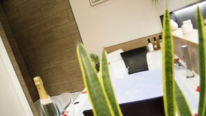 A hotel room with a wooden floor, a bed with a black headboard, a white bedside table with toiletries, and a potted plant.