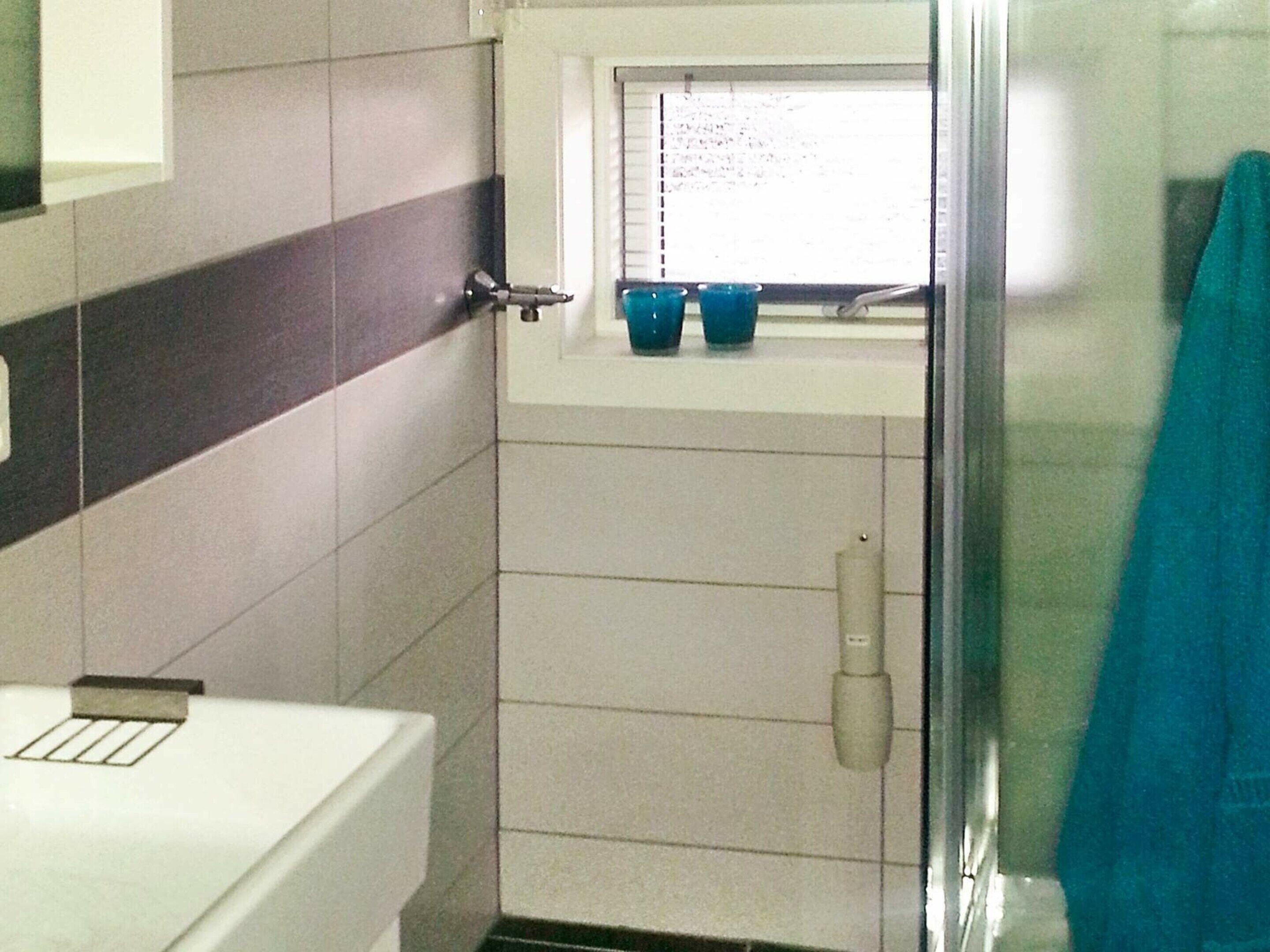 A bathroom with a toilet, a window with a shelf holding cups, and a shower area.