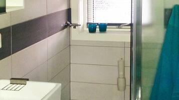 A bathroom with a toilet, a window with a shelf holding cups, and a shower area.