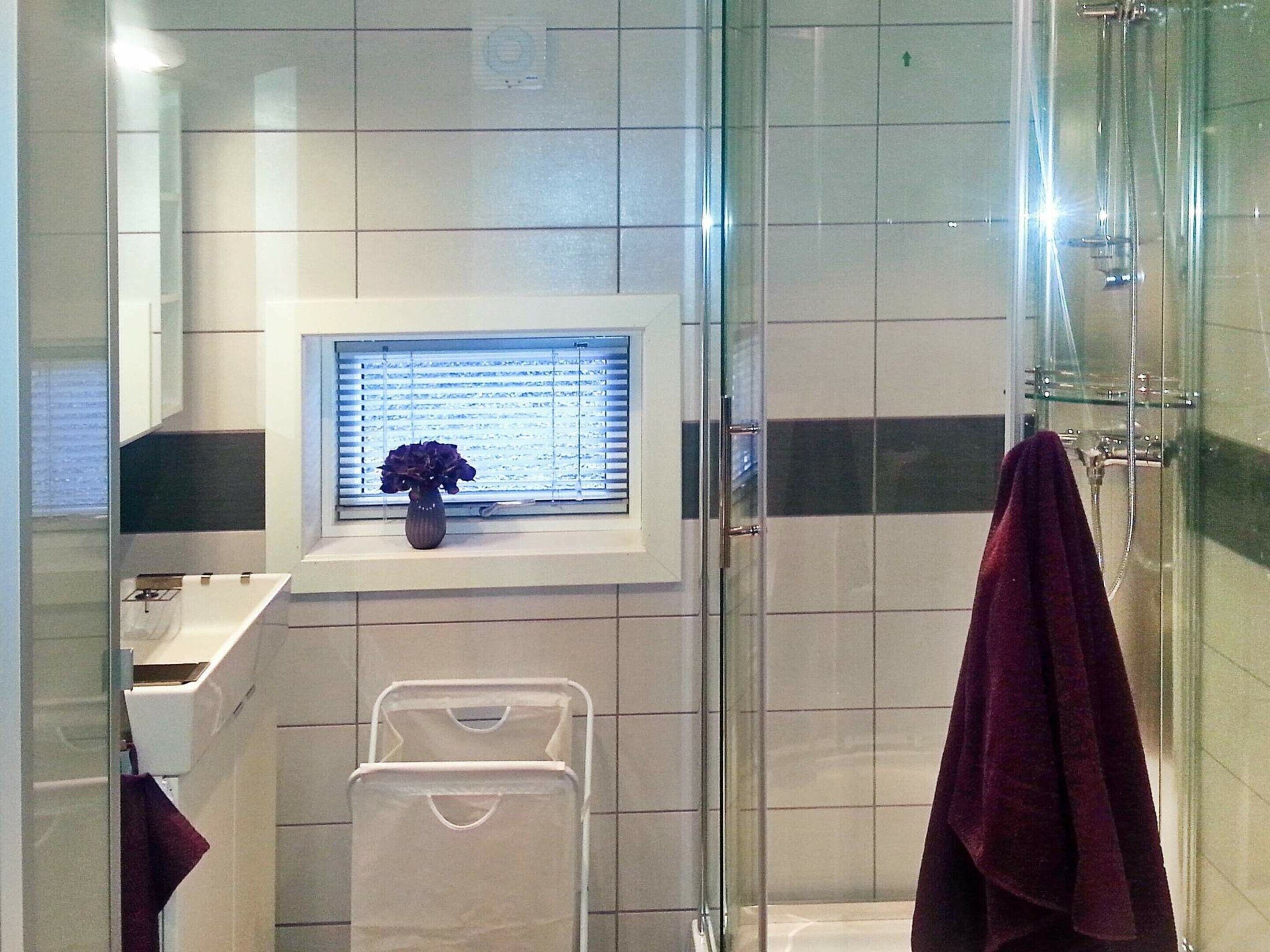 A bathroom with a glass shower enclosure, a window with blinds, a towel rack, and a small vase with flowers on the windowsill.