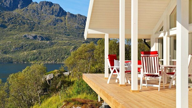 Una terraza de madera con columnas blancas y sillas a rayas rojas y blancas, con vistas a un lago y montañas.