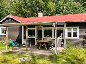 A wooden cabin with a red roof, a covered patio with a table and chairs, and a hammock.