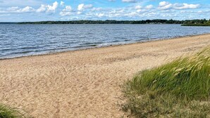 Hiekkainen ranta, jossa on tyyni järvi, ruohikkoisia alueita ja kauempana näkyvä puurivi osittain pilvisen taivaan alla.