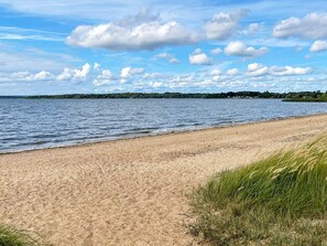 A sandy beach with a calm lake, grassy areas, and a distant tree line under a partly cloudy sky.