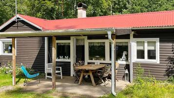 A wooden cabin with a red roof, a covered patio with a table and chairs, and a hammock.