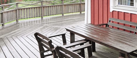 A wooden deck with a picnic table and benches, surrounded by a wooden railing, with a red building in the background.