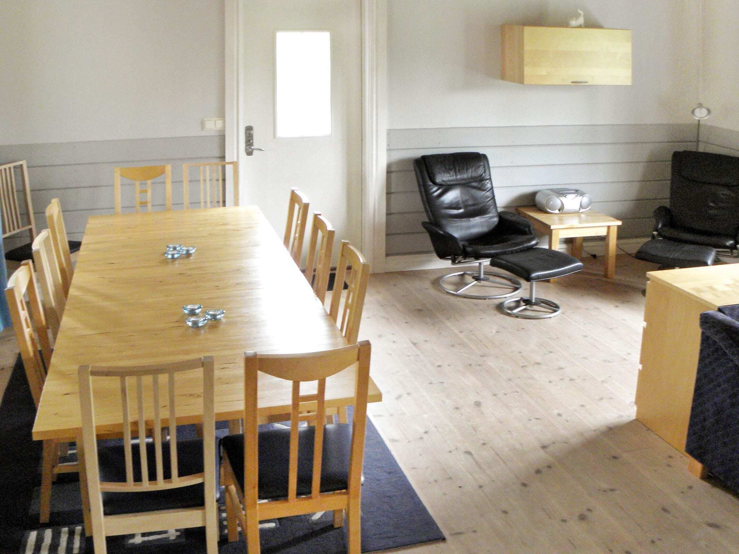 A room with a long wooden table, several chairs, a black leather chair, a small table with a speaker, and a wall-mounted shelf.
