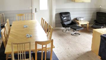 A room with a long wooden table, several chairs, a black leather chair, a small table with a speaker, and a wall-mounted shelf.