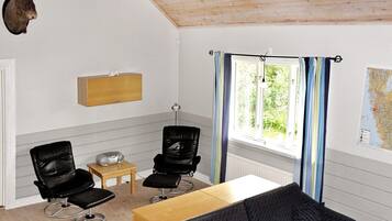 Quarto com teto de madeira, cabeça de animal montada, armário de madeira, duas cadeiras pretas, uma mesa pequena, janela com cortinas e um mapa na parede.