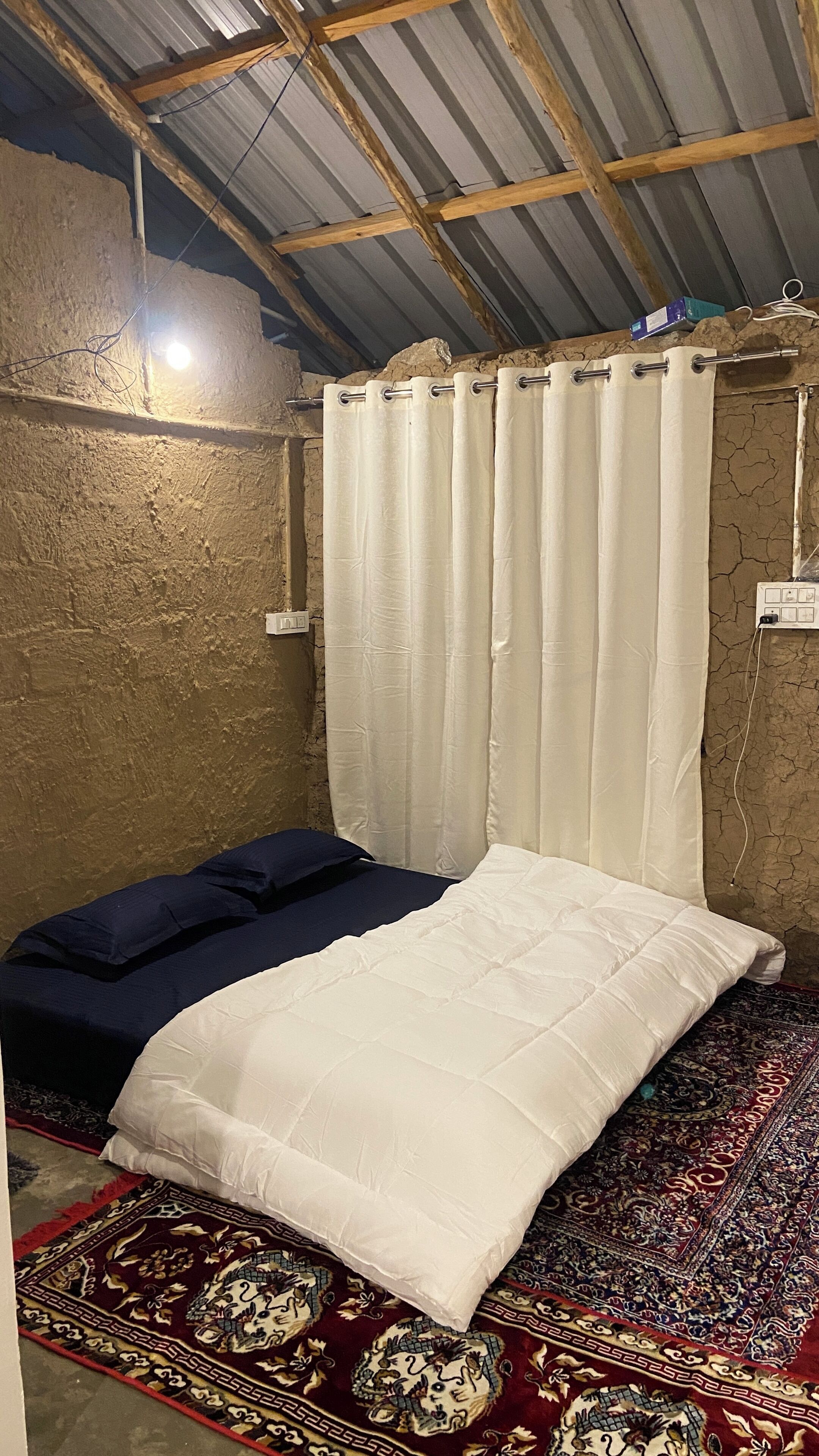 Ein Einzelbett mit weißer Matratze und blauer Decke, ein roher Teppich mit Muster sowie ein weißer Vorhang.
