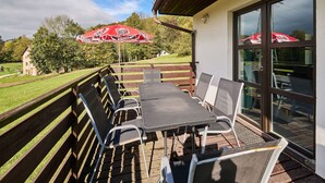 An outdoor patio with a table and chairs, a red umbrella, and a view of a grassy area with trees and a building in the background. - Green Valley Park Stárkov 1 by Interhome (Stárkov)