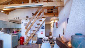 Une cuisine chaleureuse avec des poutres en bois, un coin repas avec une table dressée pour deux personnes, et un escalier menant à l’étage supérieur.