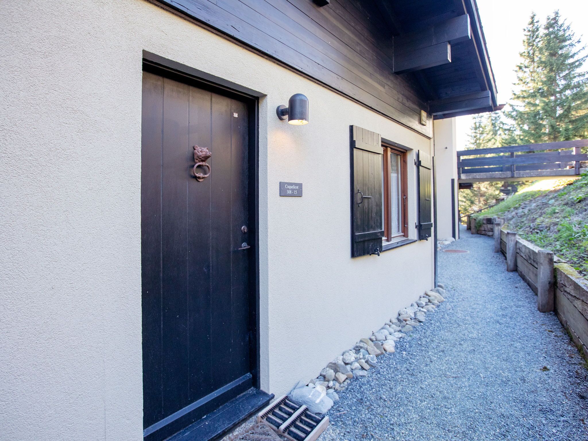 A building with a dark wooden door, a small window with shutters, and a gravel pathway leading to a wooden bridge.