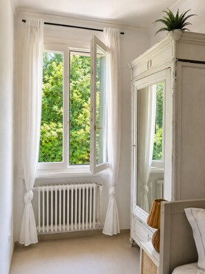 A room with a large window, white curtains, a radiator, and a white wardrobe.